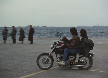 Movie still from “Landscape in the Mist” (1988), directed by Theodoros Angelopoulos – A group of people riding a motorcycle near the ocean; Wide shot, High angle