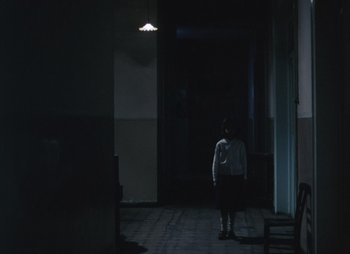 Movie still from “Landscape in the Mist” (1988), directed by Theodoros Angelopoulos – A person standing in a dark room under a light; Wide shot, High angle