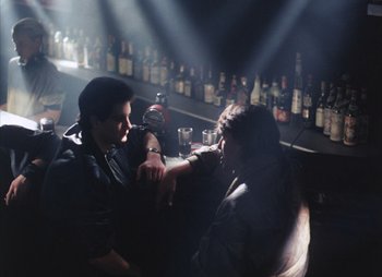 Movie still from “Landscape in the Mist” (1988), directed by Theodoros Angelopoulos – Two people sitting at a bar with bottles of alcohol behind them; Wide shot, Over the shoulder angle