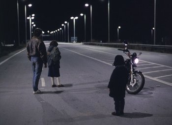 Movie still from “Landscape in the Mist” (1988), directed by Theodoros Angelopoulos – Two people and a child standing on the side of a road; Wide shot, High angle