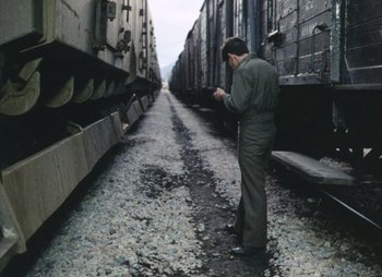Movie still from “Landscape in the Mist” (1988), directed by Theodoros Angelopoulos – A man standing on the side of a train track looking at his cell phone; Wide shot, High angle