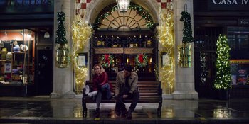 Movie still from “Last Christmas” (2019), directed by Paul Feig – A man and a woman sitting on a bench in front of a building; Wide shot, Low angle