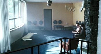 Movie still from “Last Night” (1998), directed by Don McKellar – Two people sitting in a room with many speakers on the wall; Wide shot, High angle