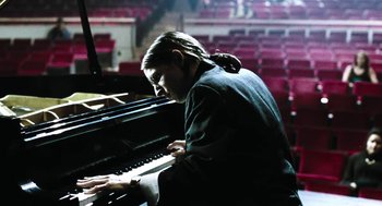 Movie still from “Last Night” (1998), directed by Don McKellar – A man sitting at a grand piano in front of an audience; Medium shot, Low angle