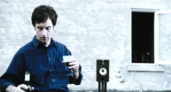 Movie still from “Last Night” (1998), directed by Don McKellar – A man sitting in front of a speaker and holding a cup; Medium shot, Low angle