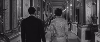 Movie still from “Last Year at Marienbad” (1961), directed by Alain Resnais – A black and white photo of people walking down a hall way; Medium shot, Over the shoulder angle