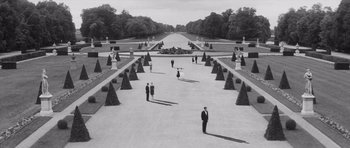 Movie still from “Last Year at Marienbad” (1961), directed by Alain Resnais – A black and white photo of people in formal wear in a formal garden; Extreme Wide shot, High angle