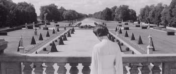 Movie still from “Last Year at Marienbad” (1961), directed by Alain Resnais – A black and white photo of a woman looking over a walkway; Wide shot, Over the shoulder angle