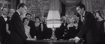 Movie still from “Last Year at Marienbad” (1961), directed by Alain Resnais – A black and white photo of a group of people standing around a lamp; Medium shot, Low angle