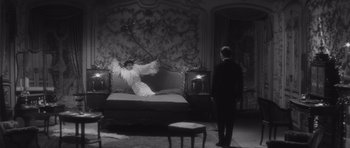 Movie still from “Last Year at Marienbad” (1961), directed by Alain Resnais – A man in a suit and a woman in a white dress on a bed in a room; Wide shot, High angle