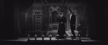 Movie still from “Last Year at Marienbad” (1961), directed by Alain Resnais – A man and a woman standing in front of candles on a stage; Wide shot, High angle