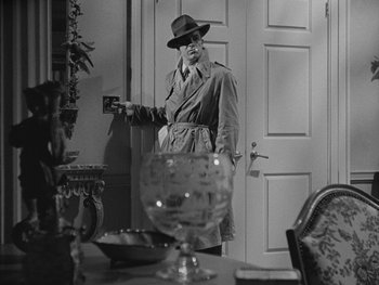 Movie still from “Laura” (1944), directed by Rouben Mamoulian – A man in a trench coat and a hat standing in front of a table; Medium shot, Over the shoulder angle
