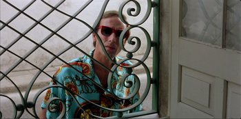 Movie still from “Law of Desire” (1987), directed by Pedro Almodóvar – A man in a blue shirt and red sunglasses looking out a window; Medium shot, Low angle