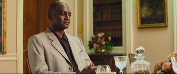 Movie still from “Layer Cake” (2004), directed by Matthew Vaughn – A man sitting at a table in front of a bookshelf; Medium shot, Over the shoulder angle