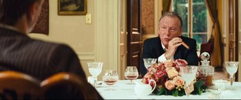 Movie still from “Layer Cake” (2004), directed by Matthew Vaughn – An older man sitting at a dinner table with wine glasses; Medium shot, Over the shoulder angle