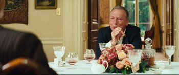Movie still from “Layer Cake” (2004), directed by Matthew Vaughn – An older man sitting at a table with a glass of wine; Medium shot, Over the shoulder angle