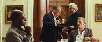 Movie still from “Layer Cake” (2004), directed by Matthew Vaughn – A man in a black jacket is signing a book; Medium shot, Over the shoulder angle