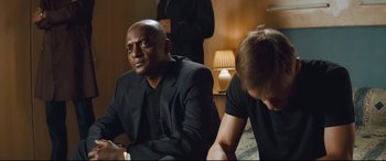 Movie still from “Layer Cake” (2004), directed by Matthew Vaughn – A man in a black suit sitting next to another man in a black suit; Medium shot, Over the shoulder angle