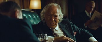 Movie still from “Layer Cake” (2004), directed by Matthew Vaughn – An older man sitting in a chair with a glass of wine; Close Up shot, Over the shoulder angle