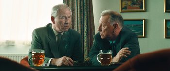 Movie still from “Layer Cake” (2004), directed by Matthew Vaughn – Two older men sitting at a table with a glass of beer in front of them; Medium shot, Over the shoulder angle
