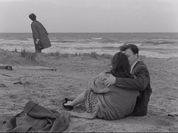 Movie still from “The Girlfriends” (1955), directed by Michelangelo Antonioni – Two people sitting on the beach with a man standing in the background; Wide shot, High angle