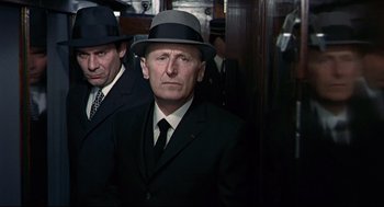 Movie still from “The Red Circle” (1970), directed by Jean-Pierre Melville – A man in a suit and a hat standing next to another man in a suit; Medium shot, Over the shoulder angle