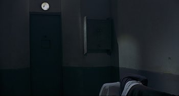 Movie still from “The Red Circle” (1970), directed by Jean-Pierre Melville – A dark room with a bed and a light on the wall behind it; Wide shot, Overhead angle