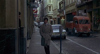 Movie still from “The Red Circle” (1970), directed by Jean-Pierre Melville – A man walking down the street in a trench coat; Wide shot, Low angle