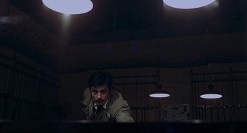 Movie still from “The Red Circle” (1970), directed by Jean-Pierre Melville – A man wearing a trench coat is sitting at a desk; Medium shot, Low angle
