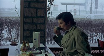 Movie still from “The Red Circle” (1970), directed by Jean-Pierre Melville – A man sitting at a table drinking from a cup; Medium shot, Over the shoulder angle