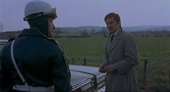 Movie still from “The Red Circle” (1970), directed by Jean-Pierre Melville – A man in a trench coat talking to another man in a police uniform; Medium shot, Over the shoulder angle
