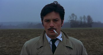 Movie still from “The Red Circle” (1970), directed by Jean-Pierre Melville – A man in a trench coat smoking a cigarette in a field; Close Up shot, High angle