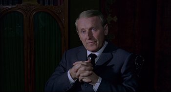 Movie still from “The Red Circle” (1970), directed by Jean-Pierre Melville – An older man wearing a suit and tie clasping his hands; Close Up shot, Over the shoulder angle