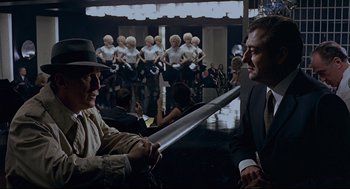 Movie still from “The Red Circle” (1970), directed by Jean-Pierre Melville – Two men are holding a sword in front of a group of people; Medium shot, Over the shoulder angle