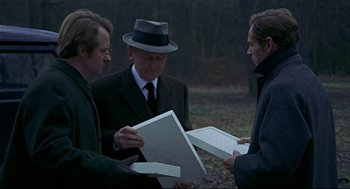 Movie still from “The Red Circle” (1970), directed by Jean-Pierre Melville – A group of men standing next to each other holding papers; Medium shot, Over the shoulder angle