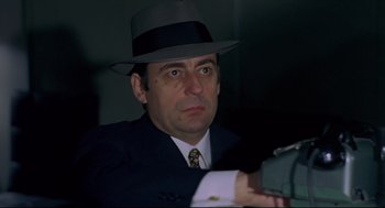 Movie still from “The Red Circle” (1970), directed by Jean-Pierre Melville – A man in a suit and a hat is holding a camera; Close Up shot, Low angle