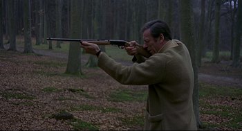 Movie still from “The Red Circle” (1970), directed by Jean-Pierre Melville – A man holding a rifle in a wooded area; Medium shot, High angle