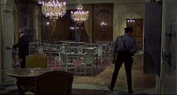 Movie still from “The Red Circle” (1970), directed by Jean-Pierre Melville – A man walking in a room filled with tables and chandeliers; Wide shot, High angle