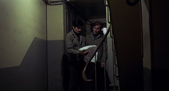Movie still from “The Red Circle” (1970), directed by Jean-Pierre Melville – Two men standing next to each other in a room; Medium shot, High angle
