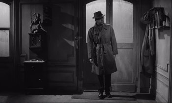 Movie still from “Le Doulos” (1962), directed by Jean-Pierre Melville – A man wearing a hat and a trench coat standing in front of an open door; Medium shot, Low angle