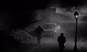 Movie still from “Le Doulos” (1962), directed by Jean-Pierre Melville – Two men are standing in the dirt near a truck; Wide shot, High angle