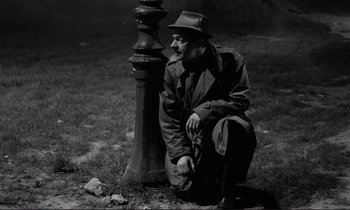 Movie still from “Le Doulos” (1962), directed by Jean-Pierre Melville – A black and white photo of a man sitting next to a fire hydrant; Medium shot, High angle