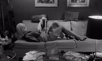Movie still from “Le Doulos” (1962), directed by Jean-Pierre Melville – A black and white photo of a woman reading on a couch; Wide shot, High angle