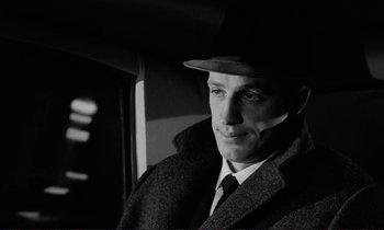 Movie still from “Le Doulos” (1962), directed by Jean-Pierre Melville – A black and white photo of a man wearing a hat and coat; Close Up shot, Over the shoulder angle