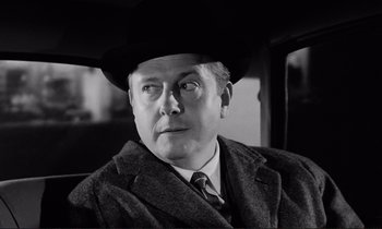 Movie still from “Le Doulos” (1962), directed by Jean-Pierre Melville – A man wearing a suit and a hat in a black and white photo; Close Up shot, Over the shoulder angle