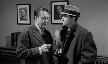 Movie still from “Le Doulos” (1962), directed by Jean-Pierre Melville – Two men in suits and a hat talking to each other in a room; Medium shot, Low angle