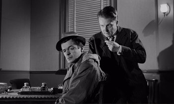 Movie still from “Le Doulos” (1962), directed by Jean-Pierre Melville – Two men in a room one of which is sitting and one of which is standing; Medium shot, Low angle