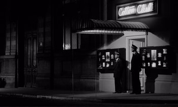 Movie still from “Le Doulos” (1962), directed by Jean-Pierre Melville – A couple of men standing next to each other on a sidewalk; Wide shot, Low angle