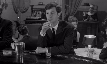 Movie still from “Le Doulos” (1962), directed by Jean-Pierre Melville – A black and white photo of a man sitting at a bar; Medium shot, Low angle