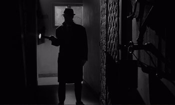 Movie still from “Le Doulos” (1962), directed by Jean-Pierre Melville – A man in a hat and a trench coat holding a flashlight; Wide shot, Low angle